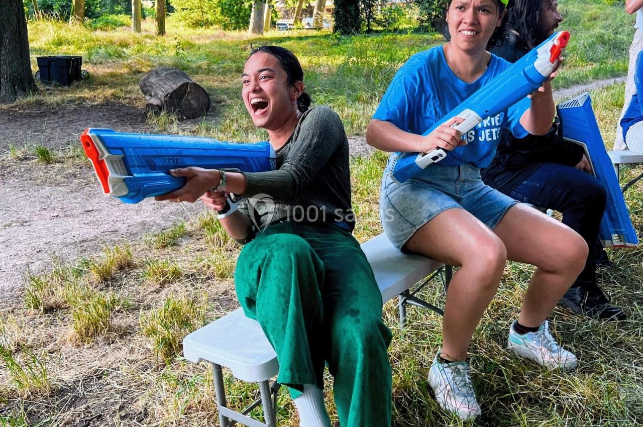 Deux personnes assises sur un banc s'amusent avec des pistolets à eau dans un parc verdoyant.