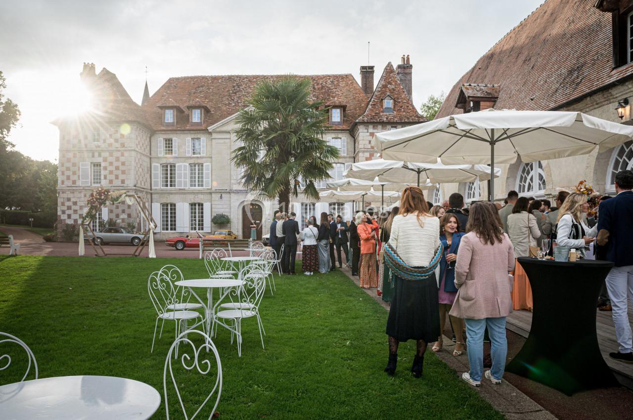 Groupe de personnes rassemblées dans un jardin devant un bâtiment historique, avec tables et parasols installés.