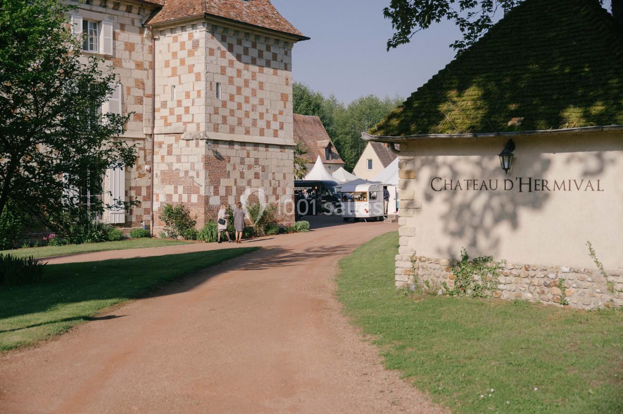 Allée bordée de pelouses menant à des bâtiments en pierre, dont un avec l'inscription ’Château d'Hermival’.