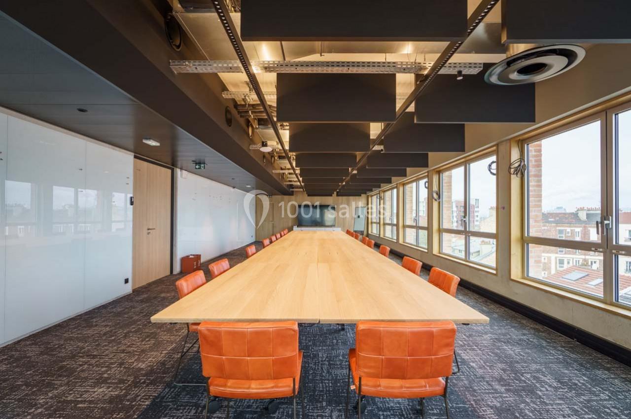 Salle de réunion moderne avec une grande table en bois, chaises orange et larges fenêtres offrant une vue sur la ville.