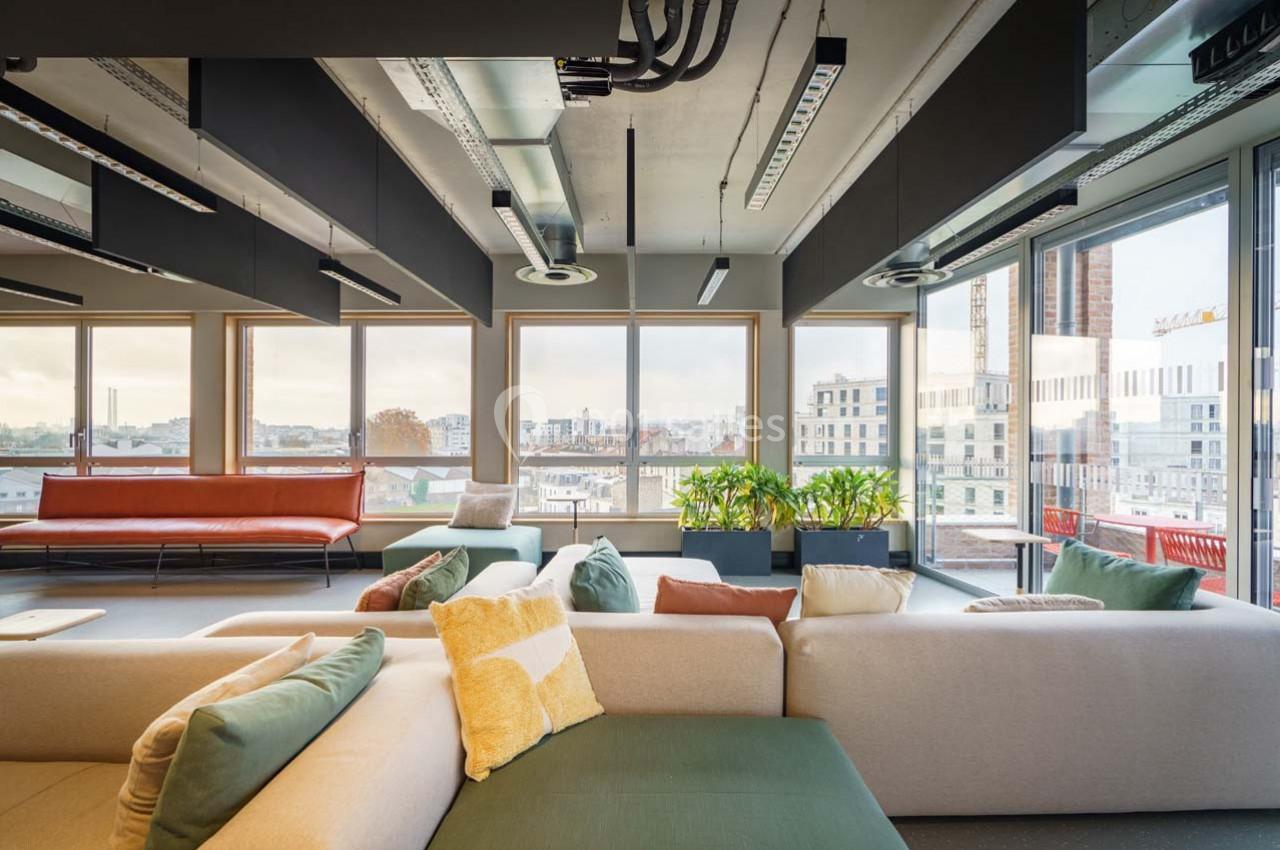 Espace lumineux avec canapés modernes, grandes fenêtres donnant sur une vue urbaine et plantes décoratives.