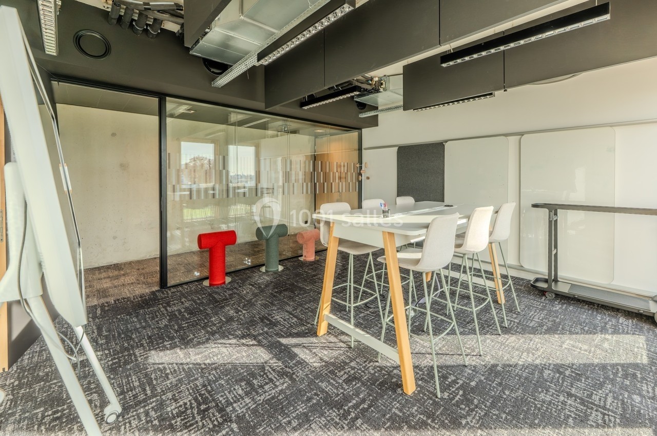 Salle de réunion moderne avec table haute, chaises, tableau blanc et cloison vitrée laissant passer la lumière naturelle.