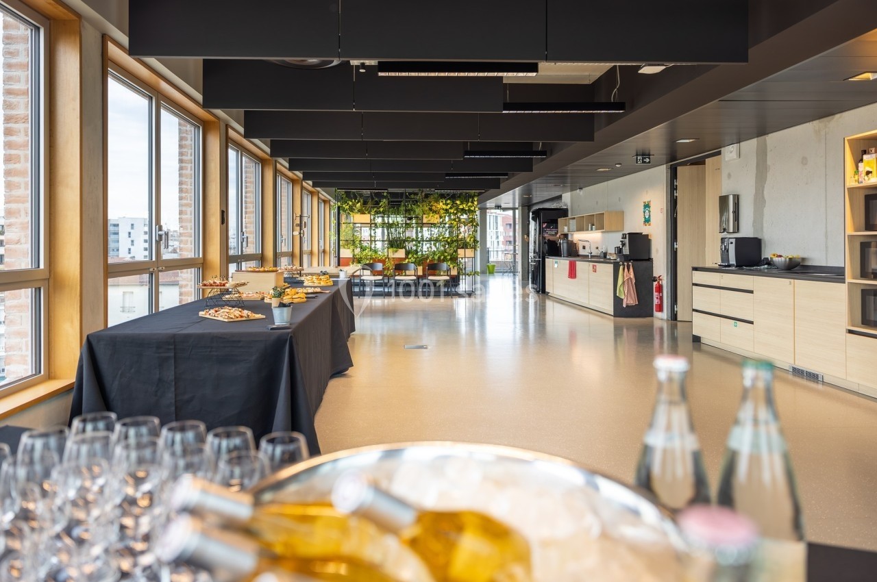 Salle lumineuse avec grandes fenêtres, tables dressées pour un buffet, et coin détente avec plantes en arrière-plan.