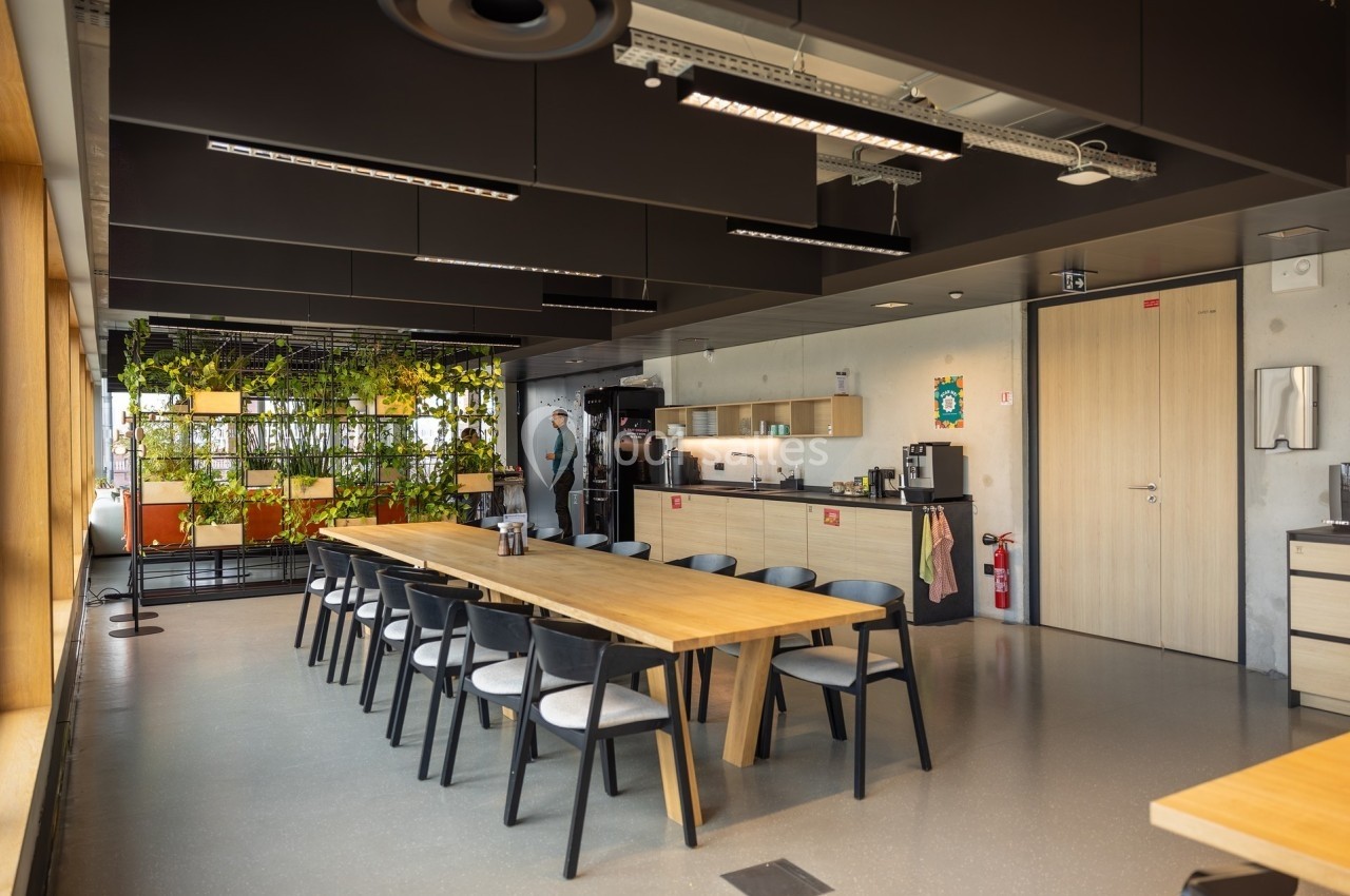 Salle de pause moderne avec tables en bois, chaises noires, kitchenette, plantes suspendues et éclairage naturel.