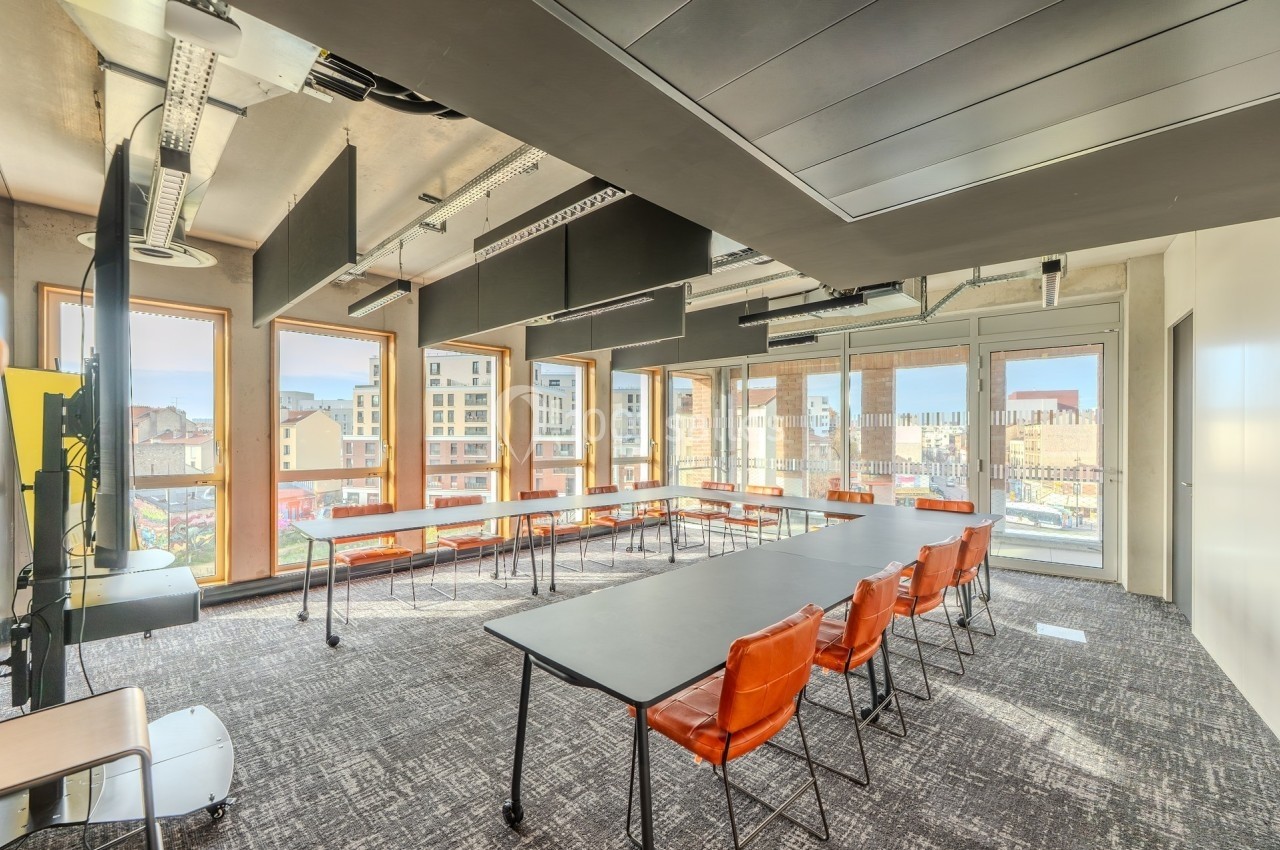 Salle de réunion lumineuse avec grandes baies vitrées, tables noires, chaises en cuir orange et vue sur des bâtiments…