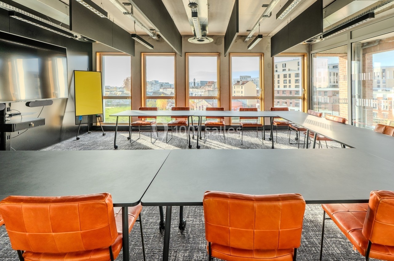 Salle de réunion lumineuse avec grandes fenêtres, tables noires, chaises orange et un tableau blanc.