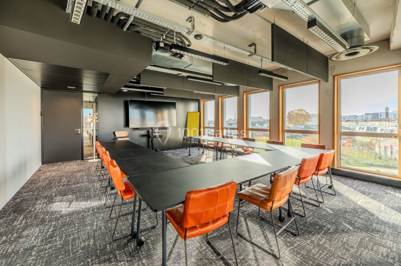Salle de réunion moderne avec grande table noire, chaises orange, écran mural et fenêtres offrant une vue extérieure.