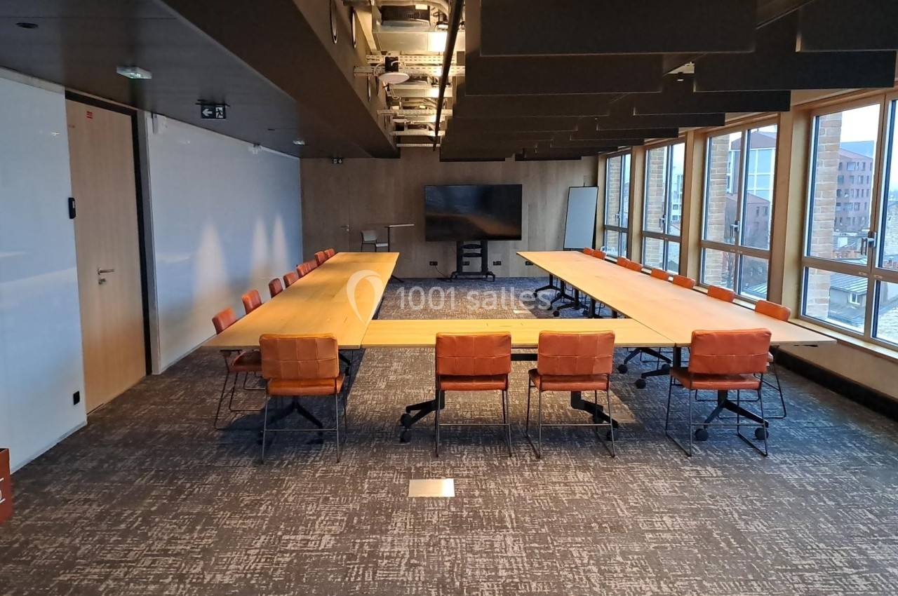 Salle de réunion moderne avec deux grandes tables en bois, chaises orange et baies vitrées donnant sur des bâtiments urbains.