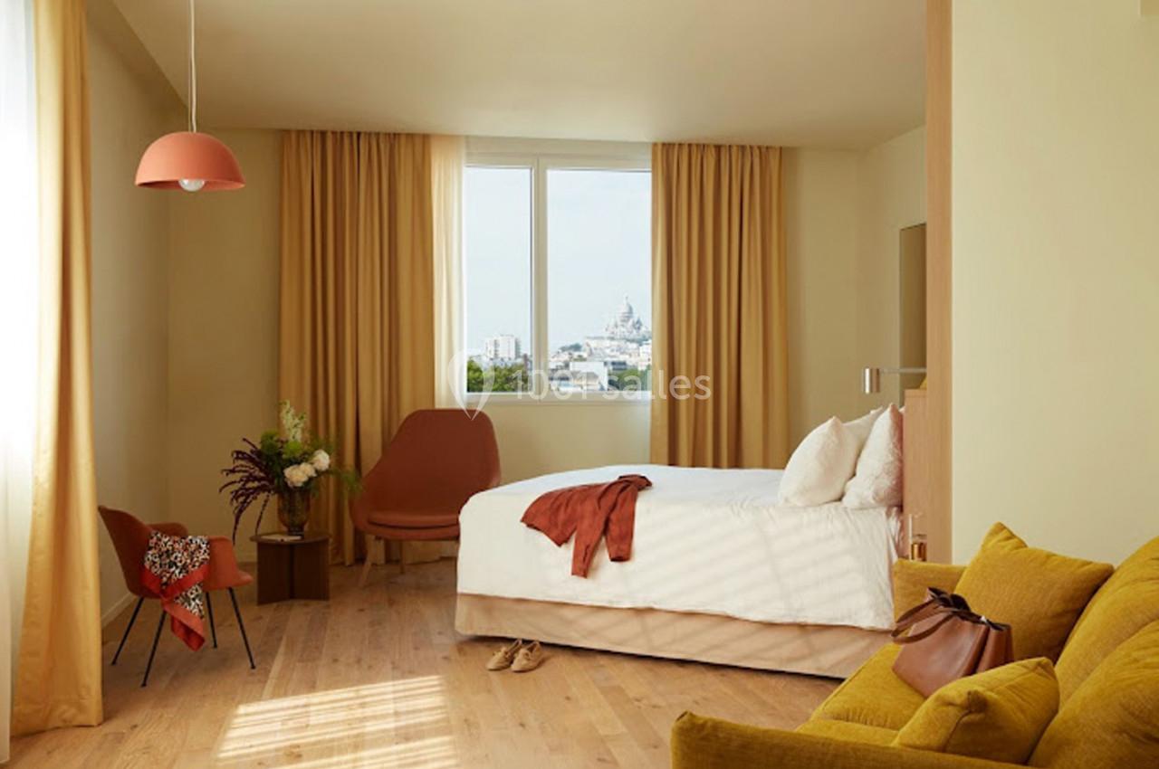 Chambre lumineuse avec lit double, fauteuils, rideaux beige, vue sur un bâtiment au loin par la fenêtre.