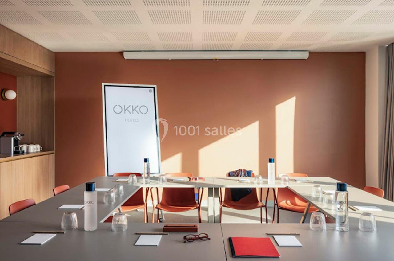 Salle de réunion lumineuse avec table en U, chaises orange, bouteilles d'eau et écran portant le logo ’Okko Hotels’.