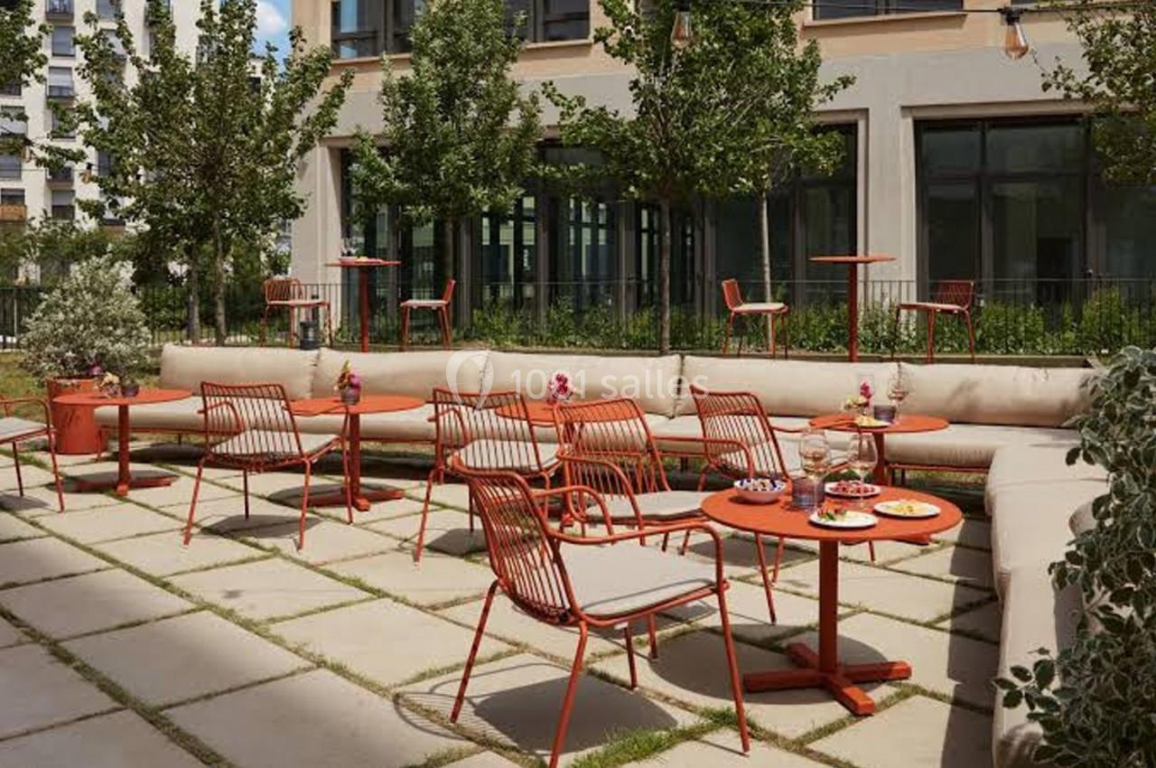Terrasse extérieure avec tables et chaises rouges, entourée de verdure et bordée par un bâtiment moderne.