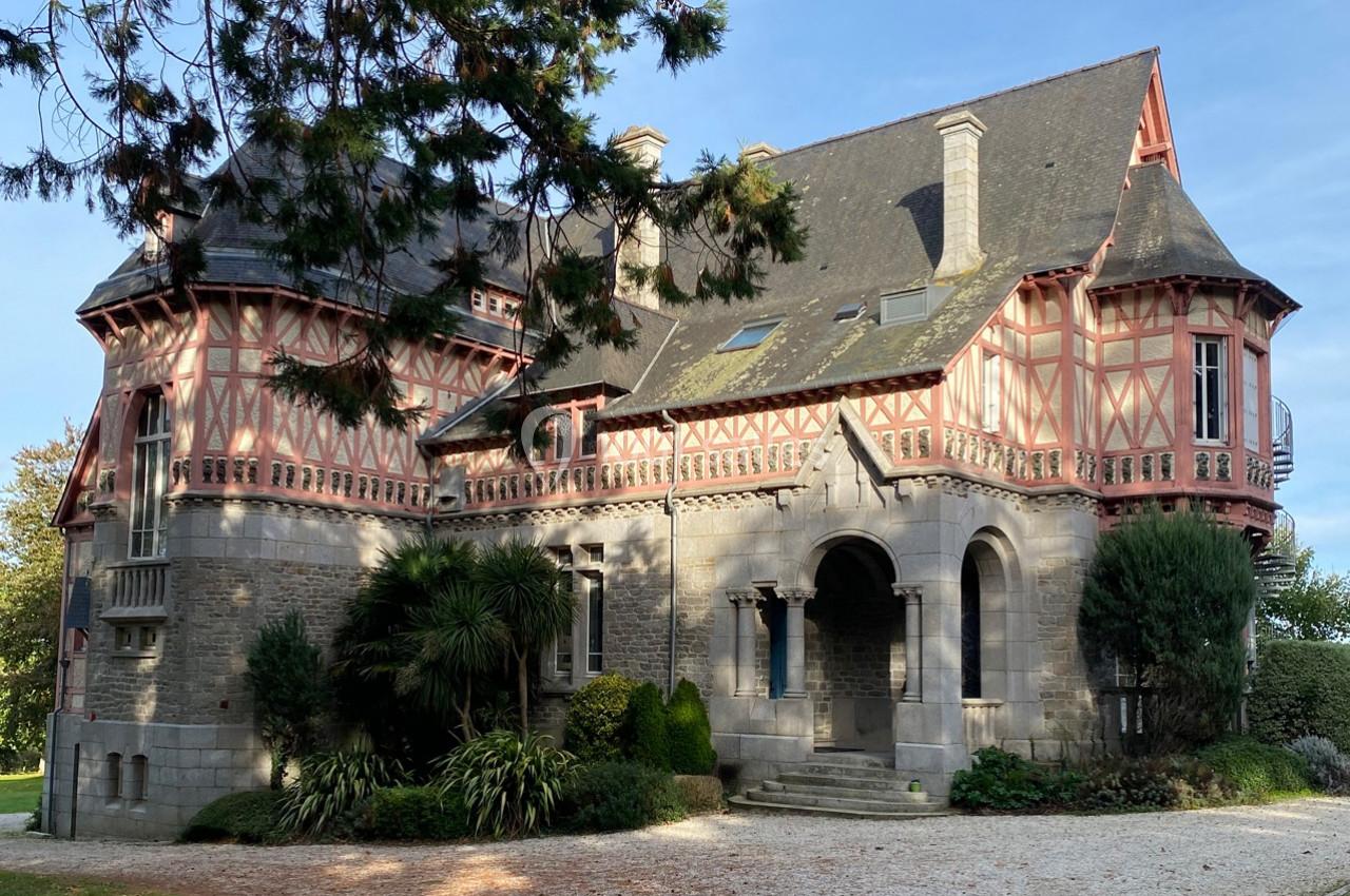Grande maison en pierre et colombages rouges, entourée de végétation, avec un ciel dégagé en arrière-plan.