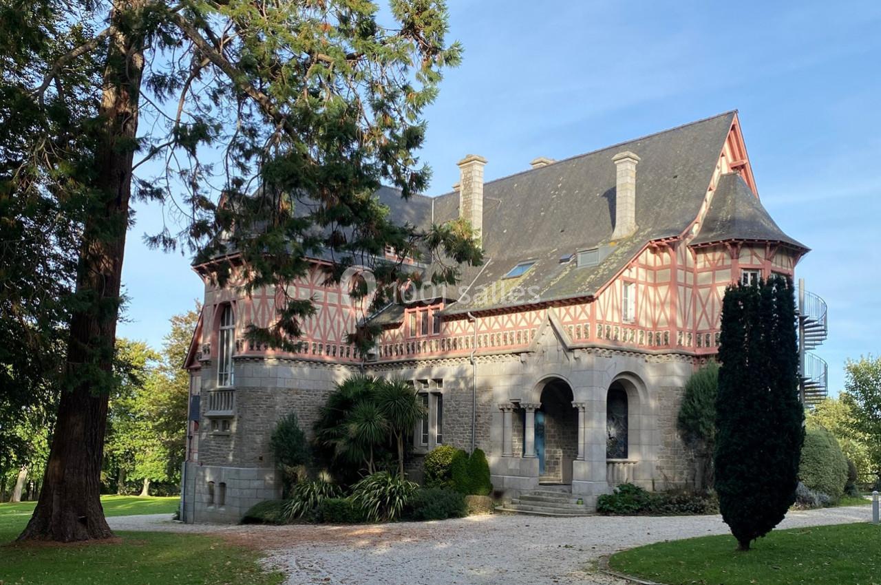 Grande maison à colombages entourée d'arbres, avec une allée gravillonnée et un ciel dégagé en arrière-plan.