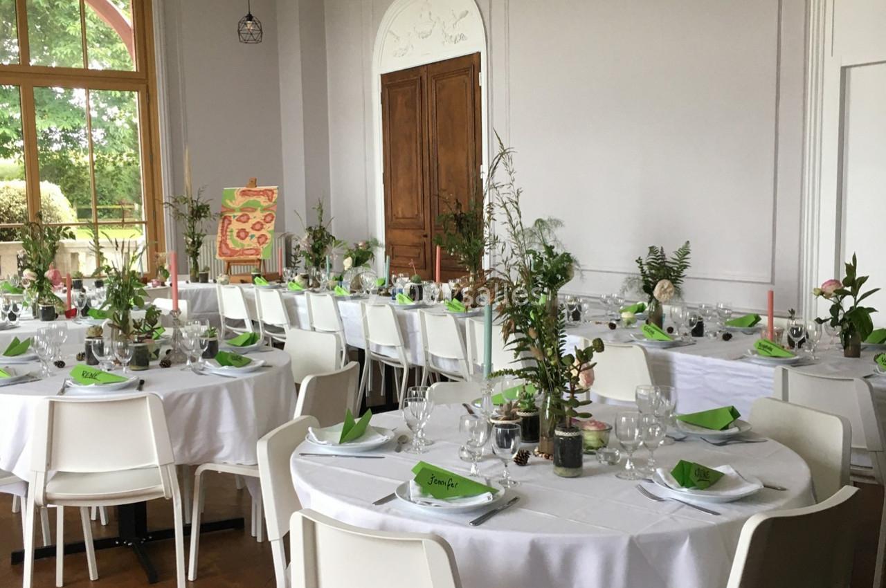 Salle de réception décorée avec des tables rondes, nappes blanches, serviettes vertes et centres de table végétaux.