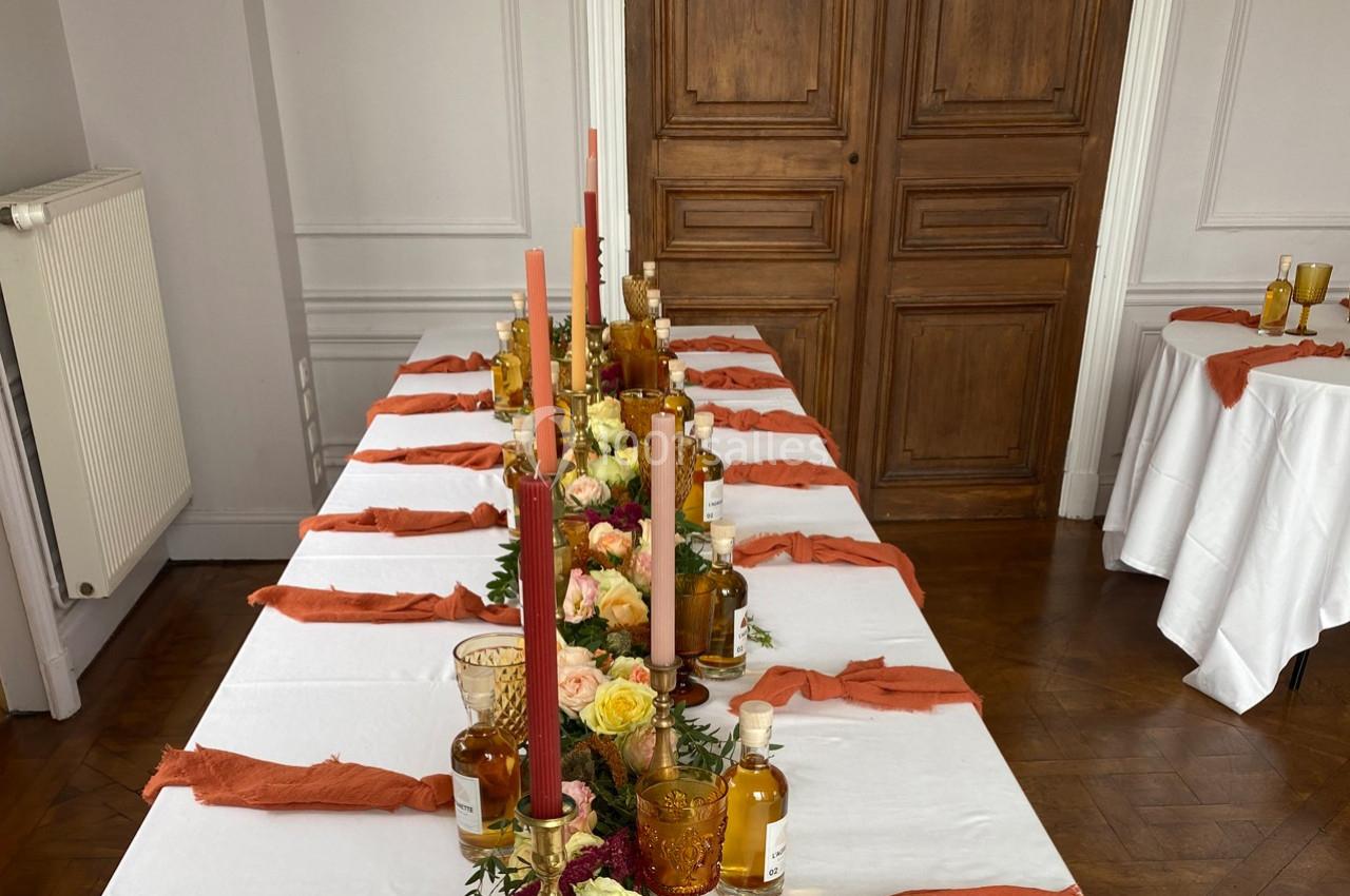 Table décorée avec des bougies colorées, des fleurs et des bouteilles ambrées, dans une pièce au parquet et porte en bois.