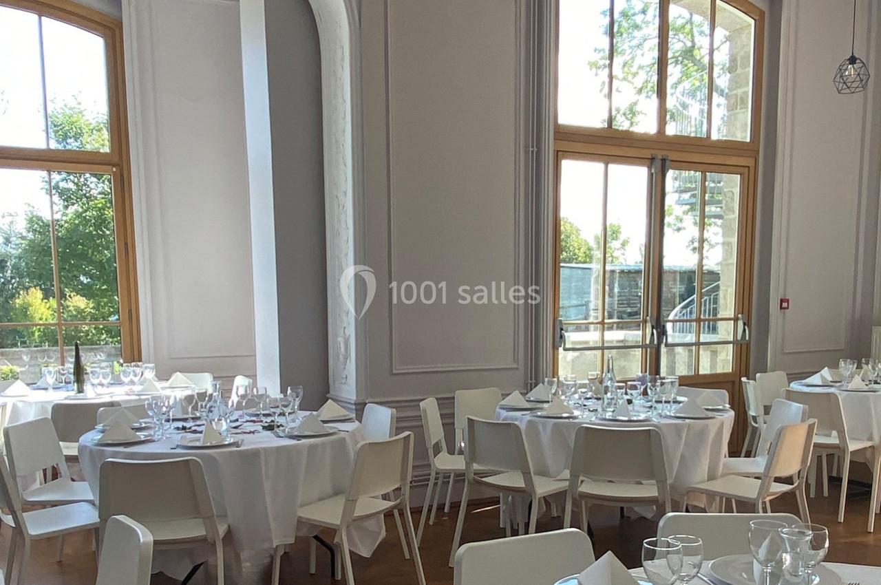 Salle lumineuse avec de grandes fenêtres, tables rondes dressées avec nappes blanches et chaises modernes.