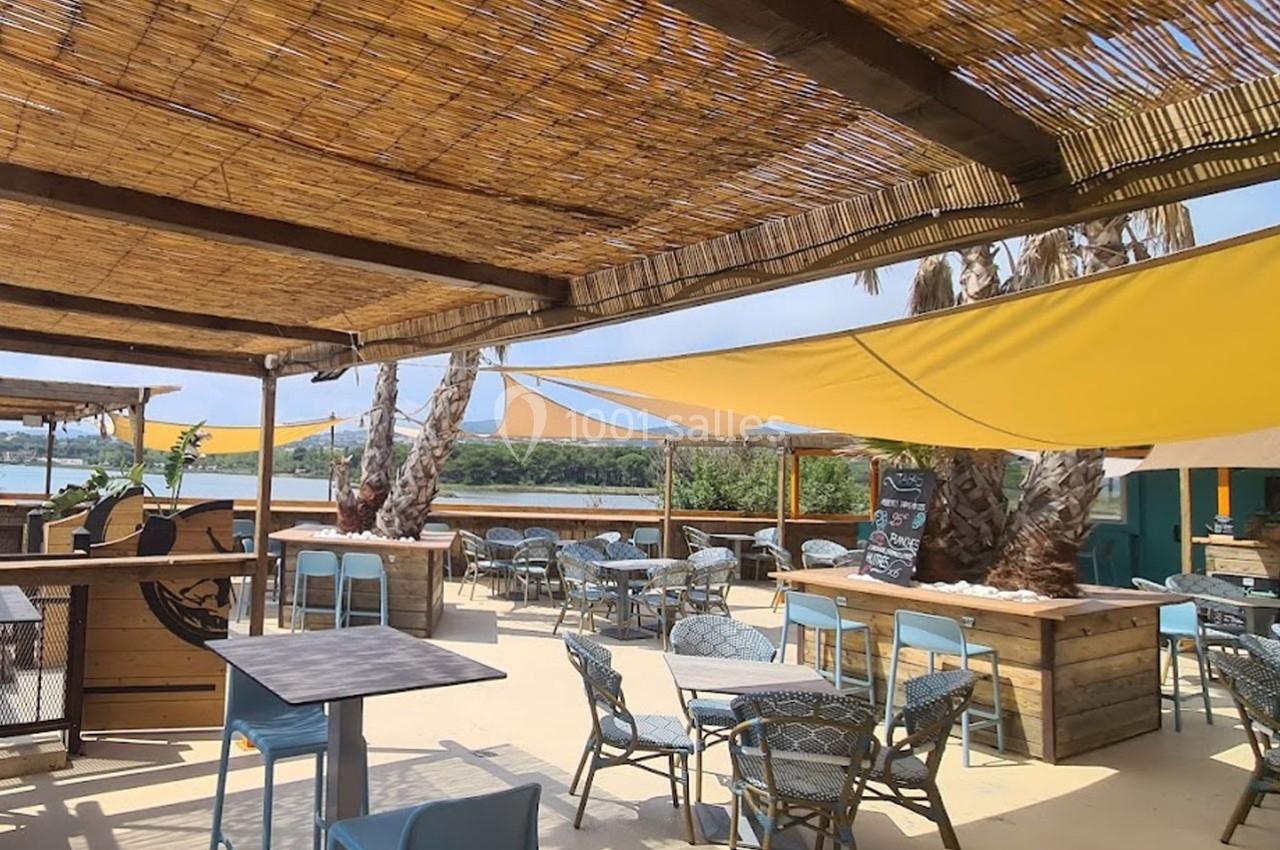 Terrasse extérieure ombragée avec tables et chaises, vue sur un plan d'eau, décorée de voiles et de palmiers.