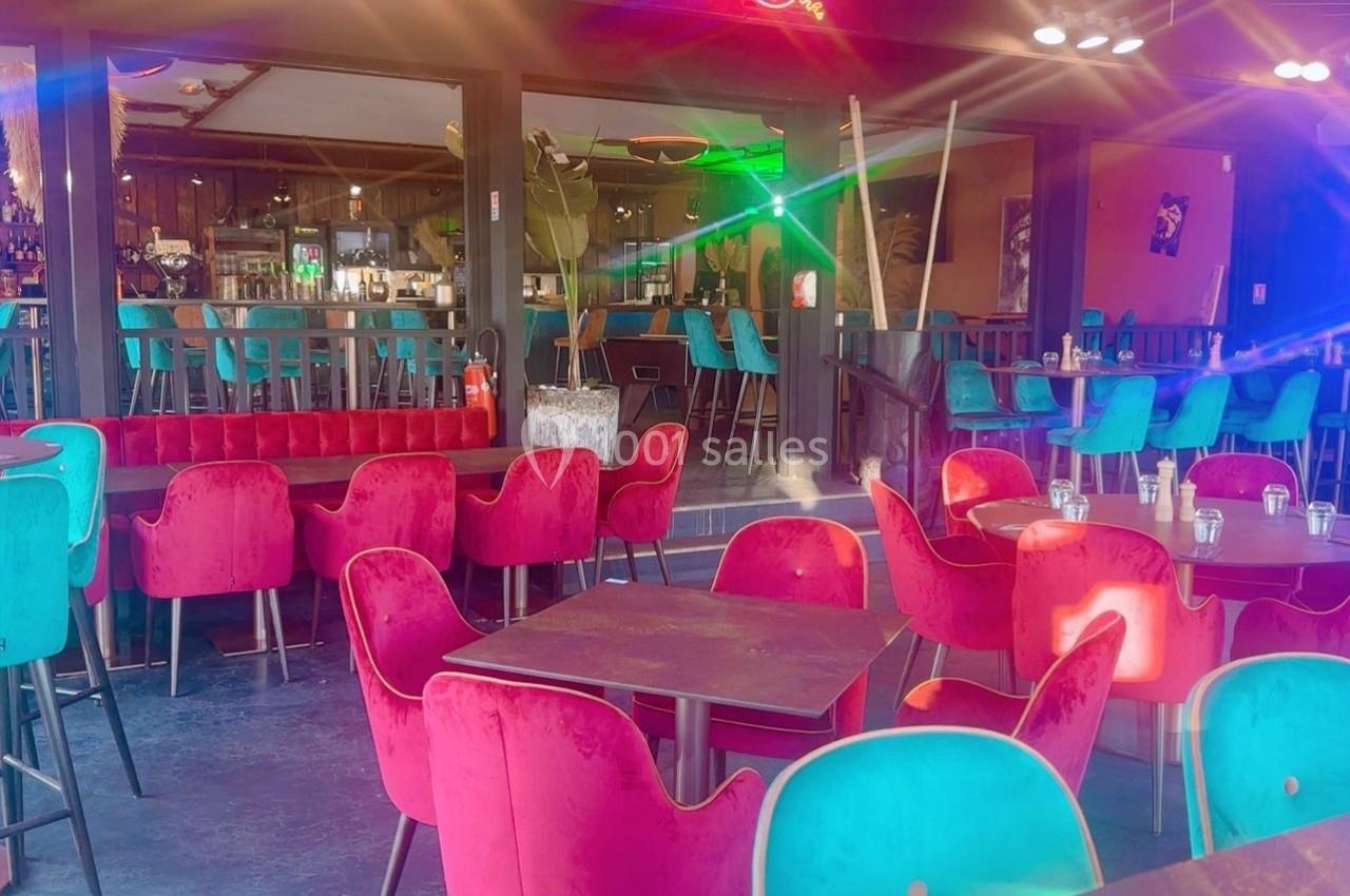 Salle de restaurant colorée avec des chaises rouges et turquoise, tables en bois et un bar en arrière-plan.