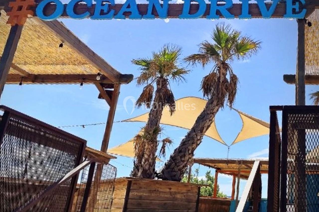 Entrée d'une terrasse en bois avec palmiers, voiles d'ombrage et une enseigne ’Ocean Drive’ en lettres bleues.