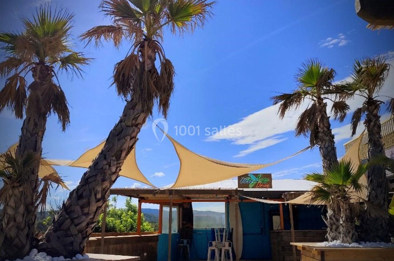 Terrasse extérieure avec palmiers, voiles d'ombrage beige et cabane en bois sous un ciel bleu.