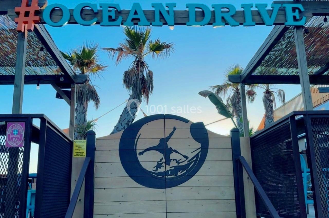Entrée en bois d'un lieu avec un logo de surfeur, palmiers en arrière-plan et enseigne ’Ocean Drive’ au sommet.