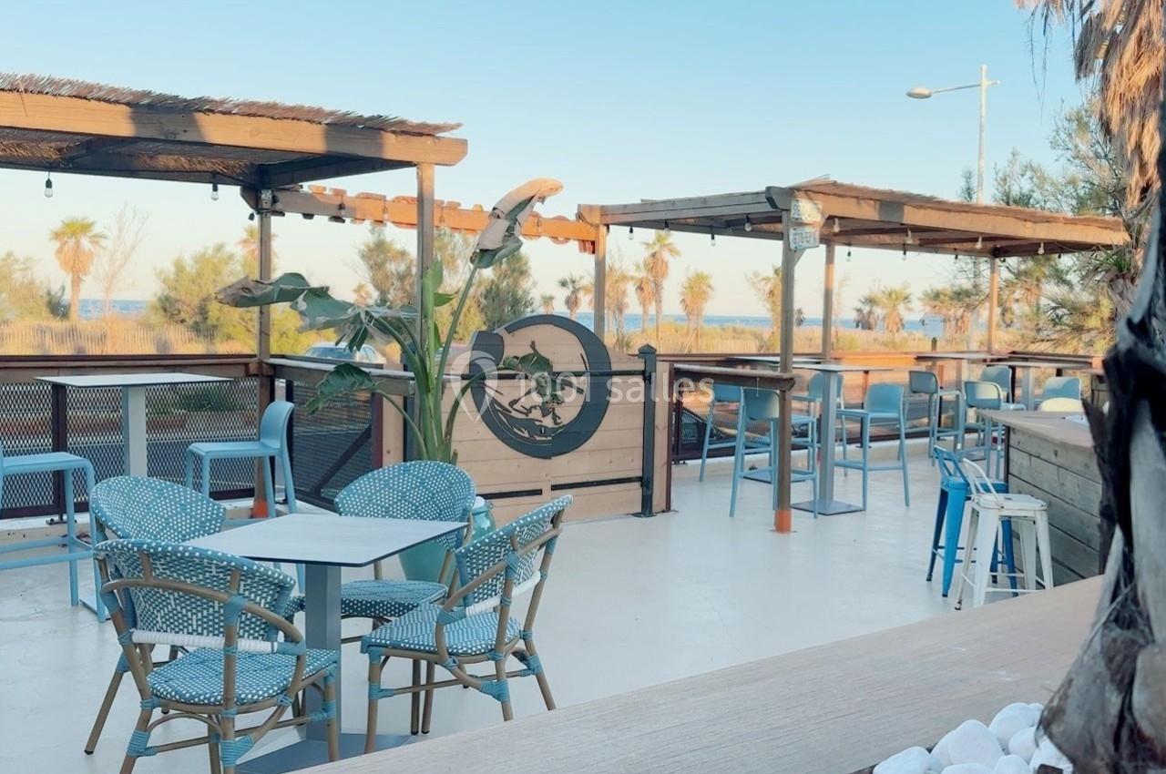 Terrasse extérieure avec tables et chaises en rotin, vue sur des arbres et la mer à l'horizon.