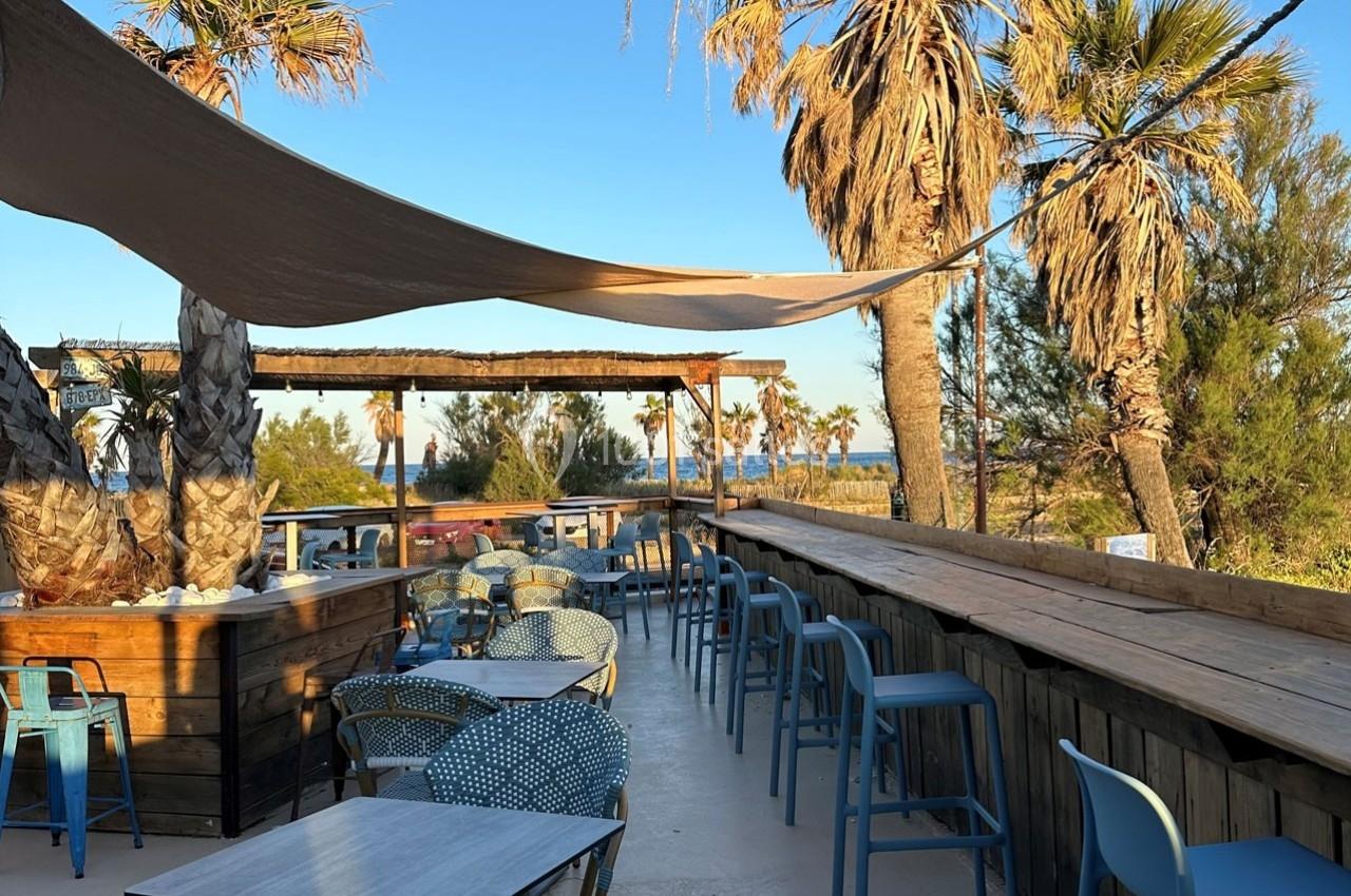 Terrasse en plein air avec tables, chaises et bar en bois, entourée de palmiers, vue sur la mer en arrière-plan.