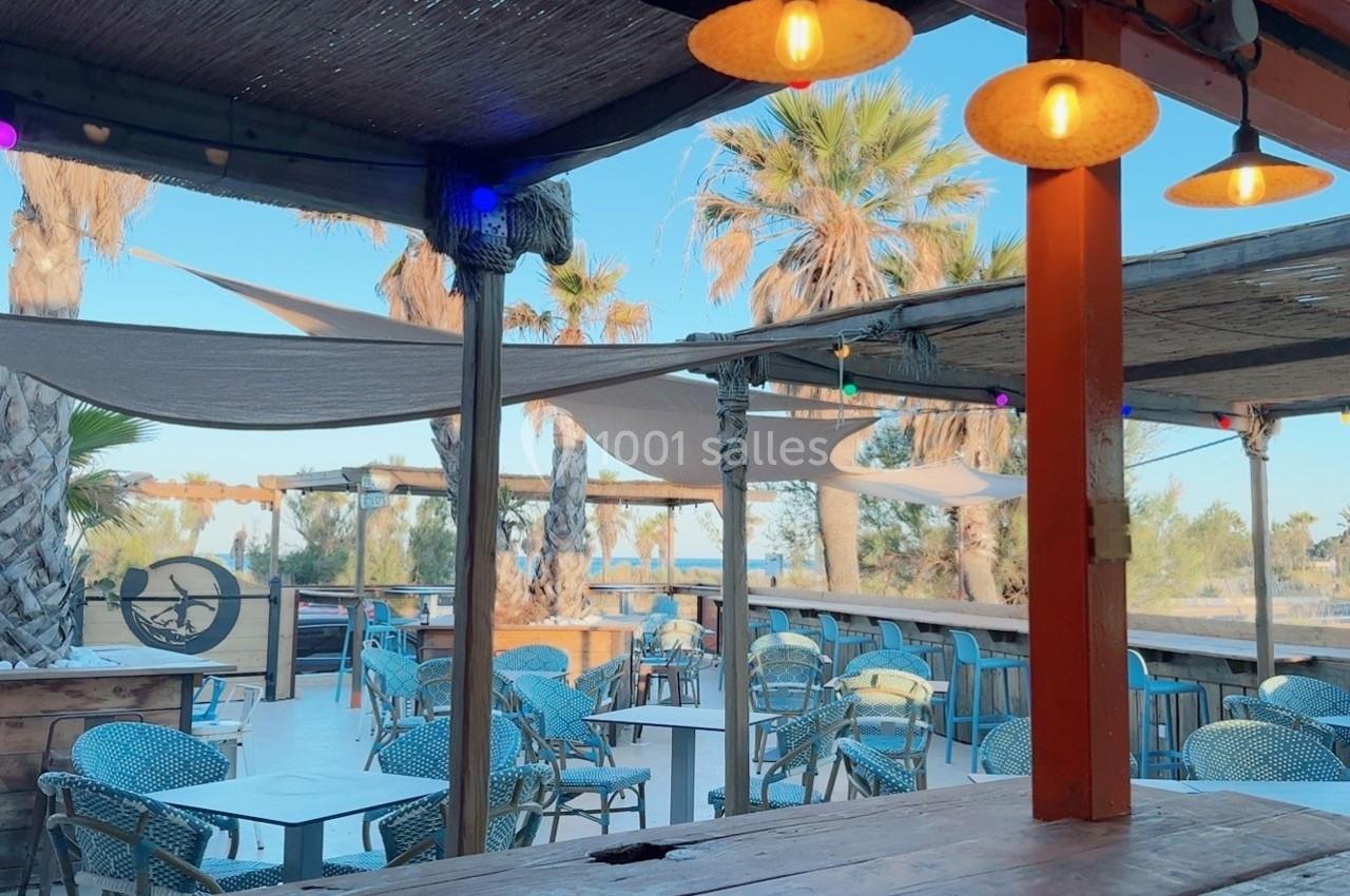 Terrasse extérieure avec tables, chaises en osier, voiles d'ombrage, palmiers et vue dégagée sur un ciel bleu.