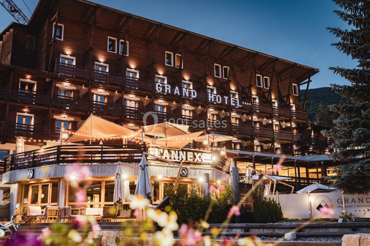 Façade éclairée d'un hôtel en bois avec terrasses et enseignes lumineuses, entouré de fleurs au premier plan.