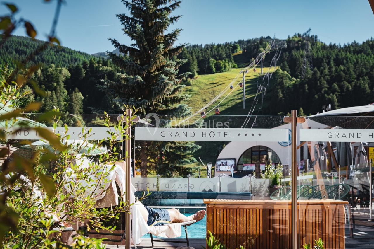 Vue d'une terrasse d'hôtel avec piscine, transat occupé, montagnes et remontées mécaniques en arrière-plan.