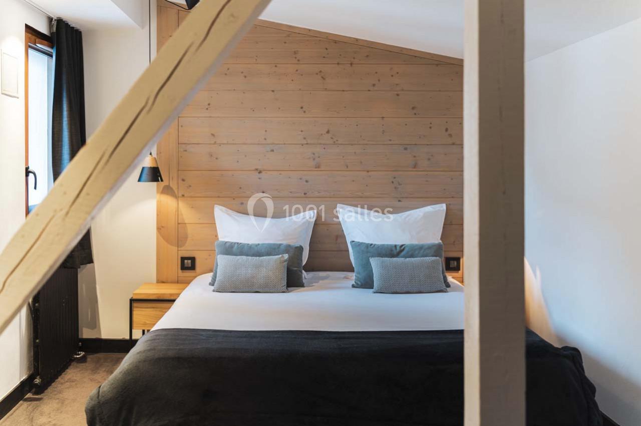Lit double avec coussins et couverture sombre dans une chambre au mur en bois clair, éclairée par une lumière naturelle.
