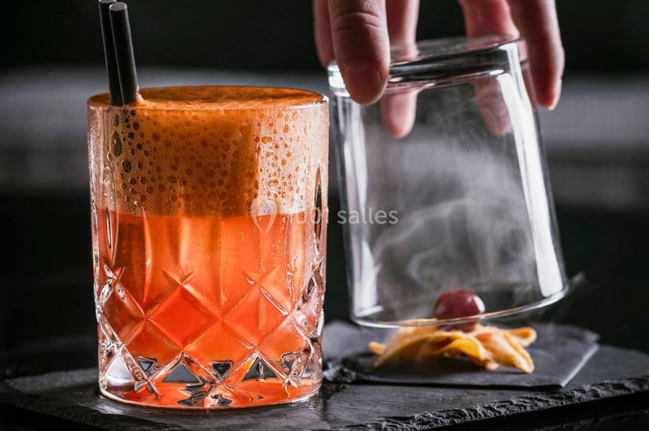 Verre de cocktail orange mousseux avec une paille, accompagné d'un verre retourné contenant une cerise et un zeste.