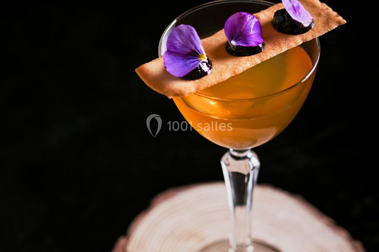 Verre à cocktail orange décoré d'une fine tranche de biscuit, de fleurs violettes et de petites garnitures noires.