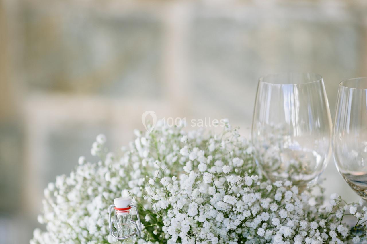 Centre de table avec gypsophile blanche, menu, bouteille de liqueur et verres à vin sur une nappe blanche.