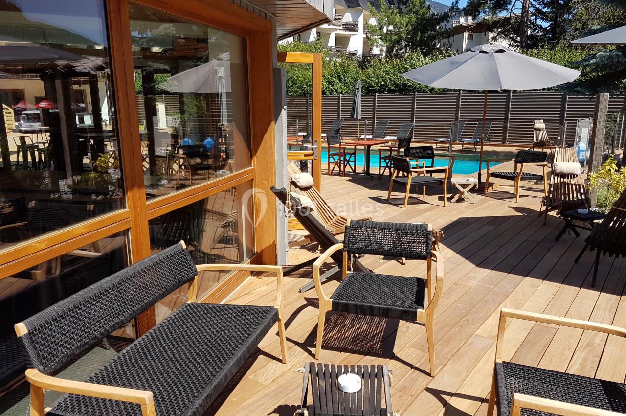 Terrasse en bois avec chaises et tables noires, parasols, et vue sur une piscine entourée de verdure.