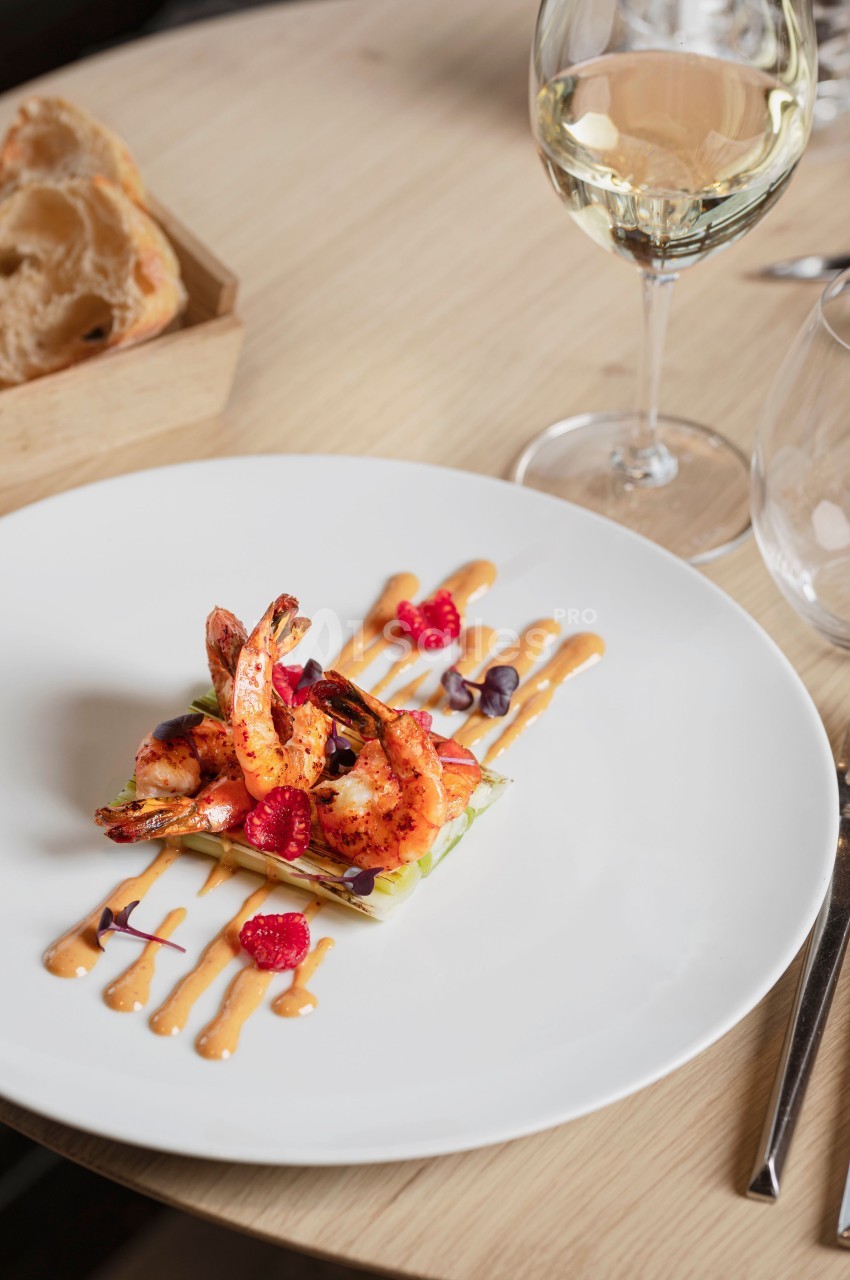 Assiette de crevettes grillées sur lit de légumes, décorée de framboises et sauce, avec verre de vin en arrière-plan.