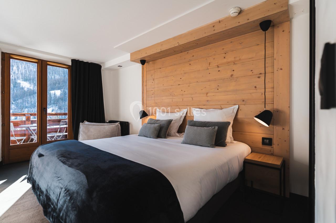 Chambre moderne avec lit double, tête de lit en bois, luminaires noirs et balcon offrant une vue sur la montagne enneigée.