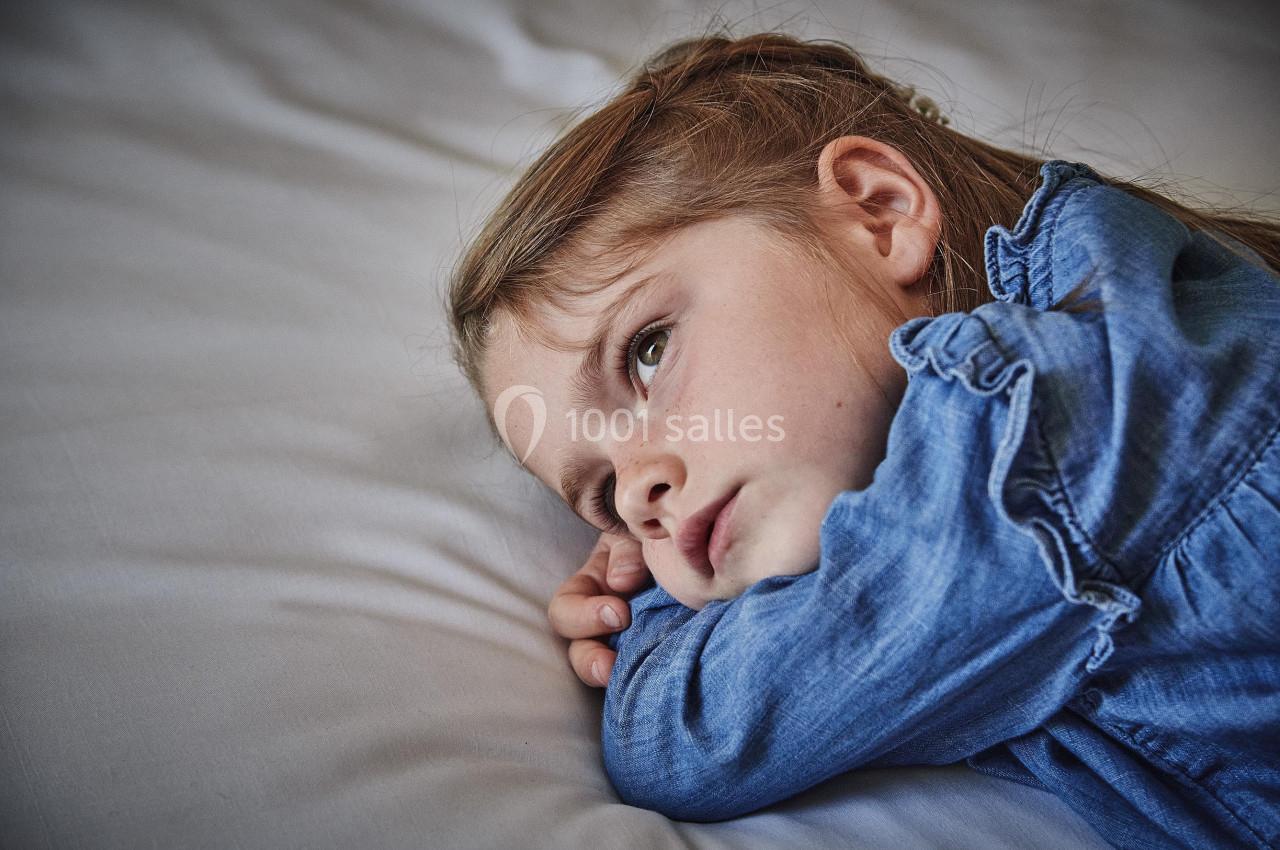 Une fillette allongée sur un lit, appuyée sur son bras, regardant au loin avec une expression pensive.