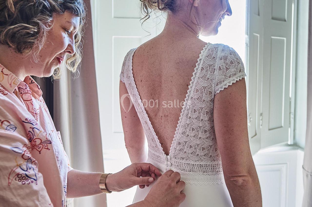 Une femme aide une mariée à ajuster sa robe blanche près d'une fenêtre lumineuse.