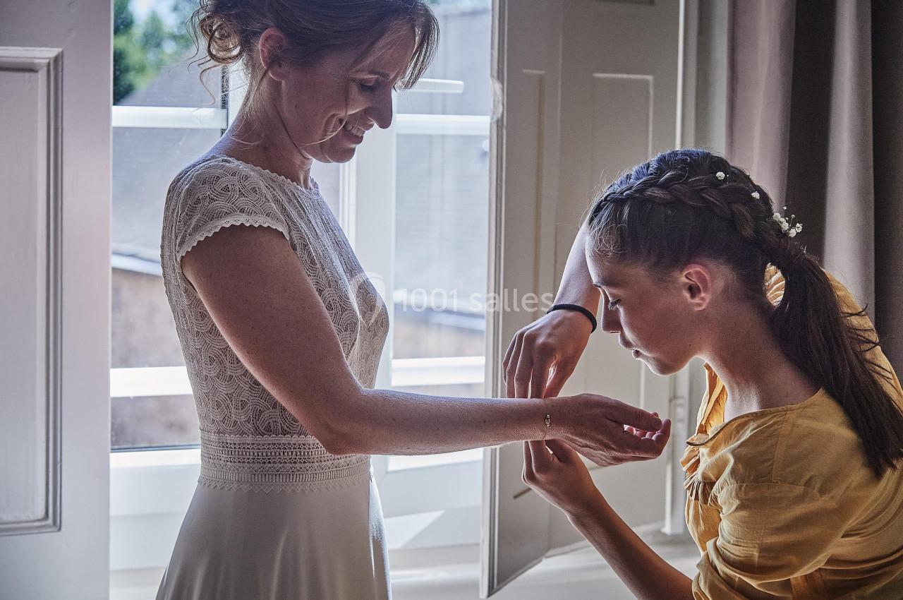 Une femme en robe blanche se fait attacher un bracelet par une jeune fille près d'une fenêtre ouverte.