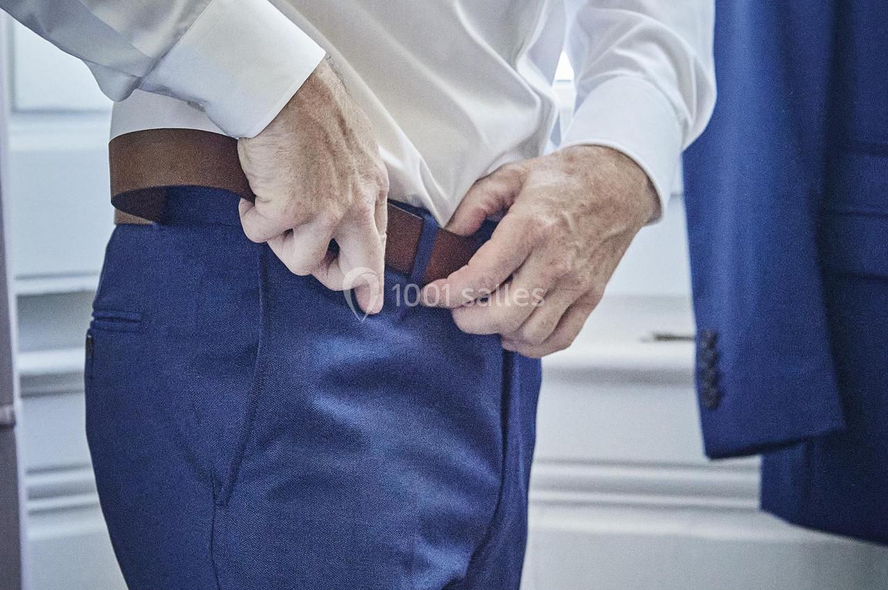 Un homme ajuste sa ceinture sur un pantalon bleu, vêtu d'une chemise blanche, près d'une veste suspendue.
