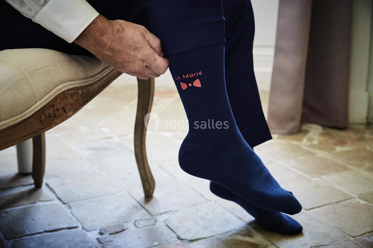 Un homme ajuste une chaussette bleue avec l'inscription ’Le Marié’ et un nœud papillon rose, assis sur une chaise.