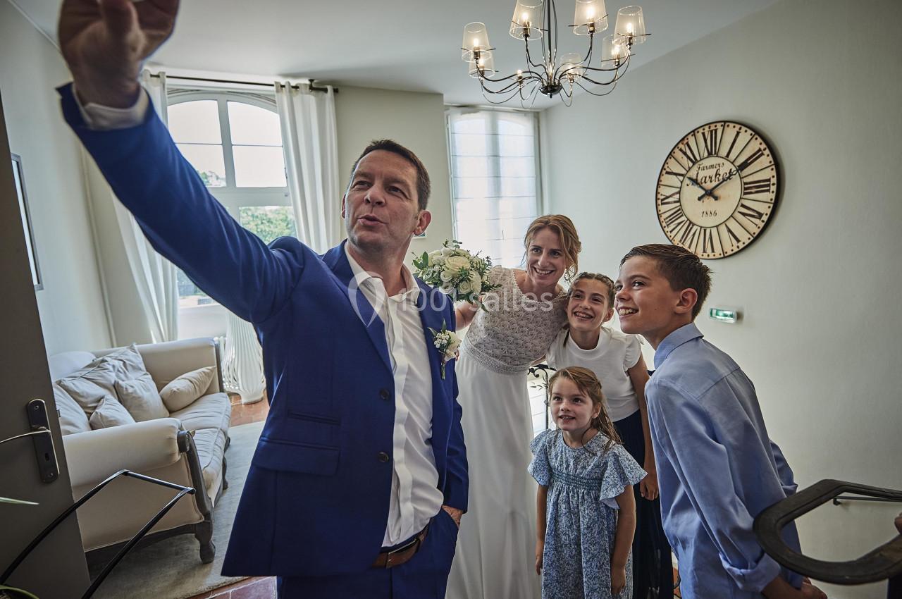 Un couple marié et trois enfants posent pour un selfie dans un salon lumineux avec une horloge murale visible.