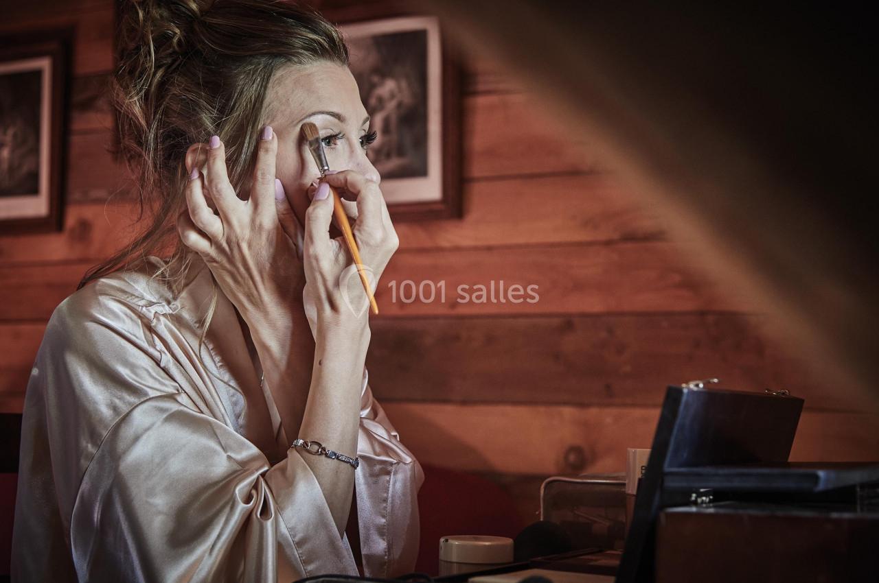 Femme appliquant du maquillage sur ses paupières devant un miroir, dans une pièce aux murs en bois.