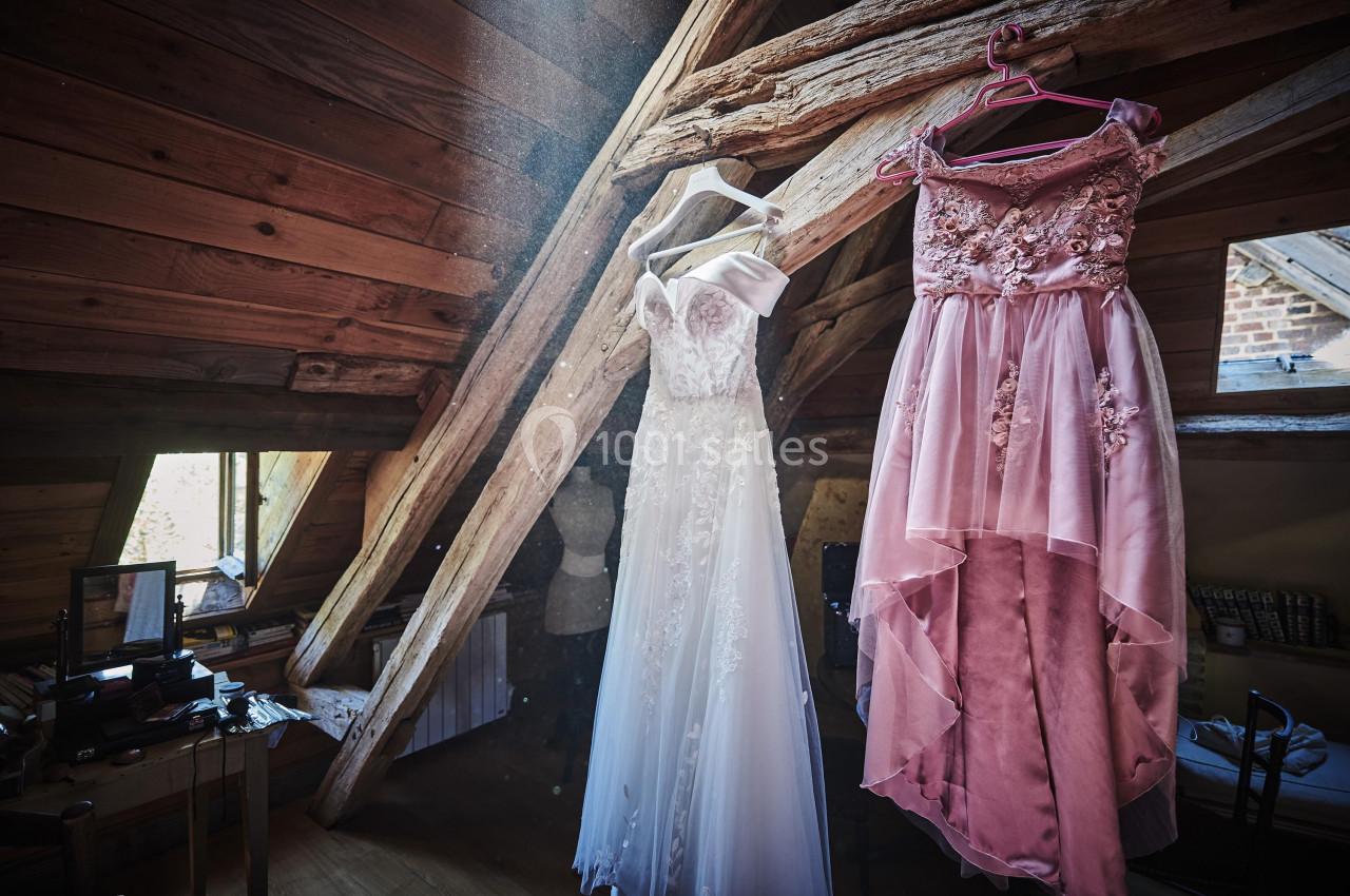 Deux robes élégantes, une blanche et une rose, suspendues dans une pièce mansardée avec des poutres apparentes.