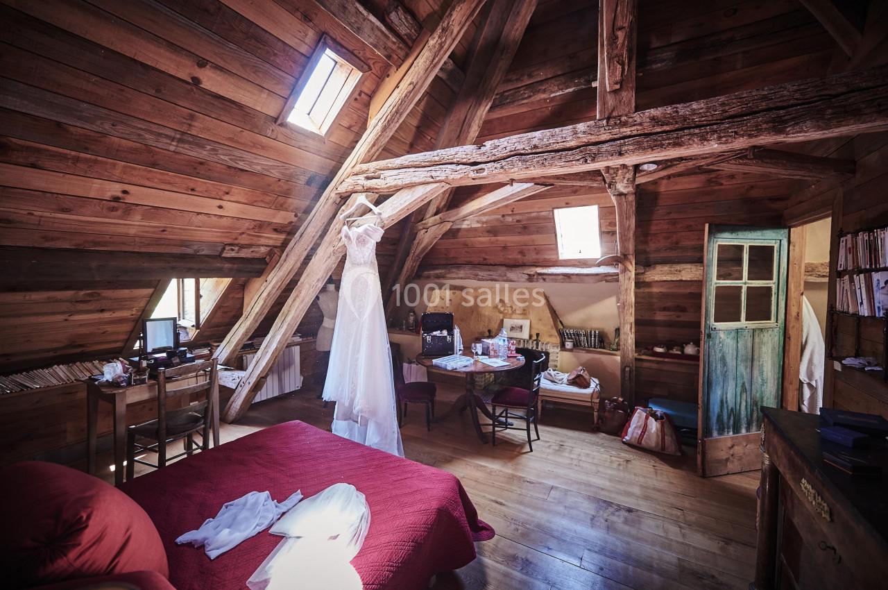 Robe de mariée suspendue dans une chambre mansardée en bois avec mobilier rustique et lumière naturelle.