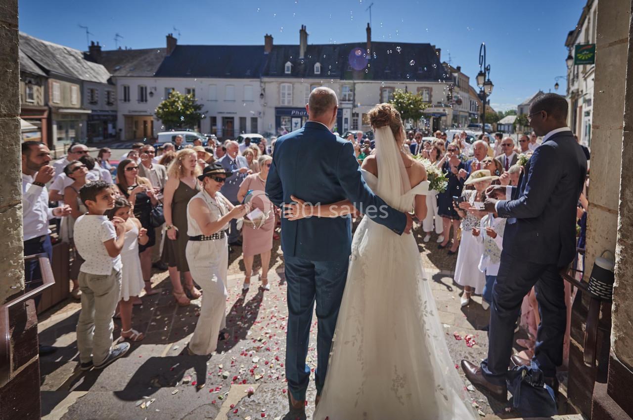 Un couple de mariés sort d'un bâtiment, entouré d'invités célébrant sous un ciel ensoleillé.