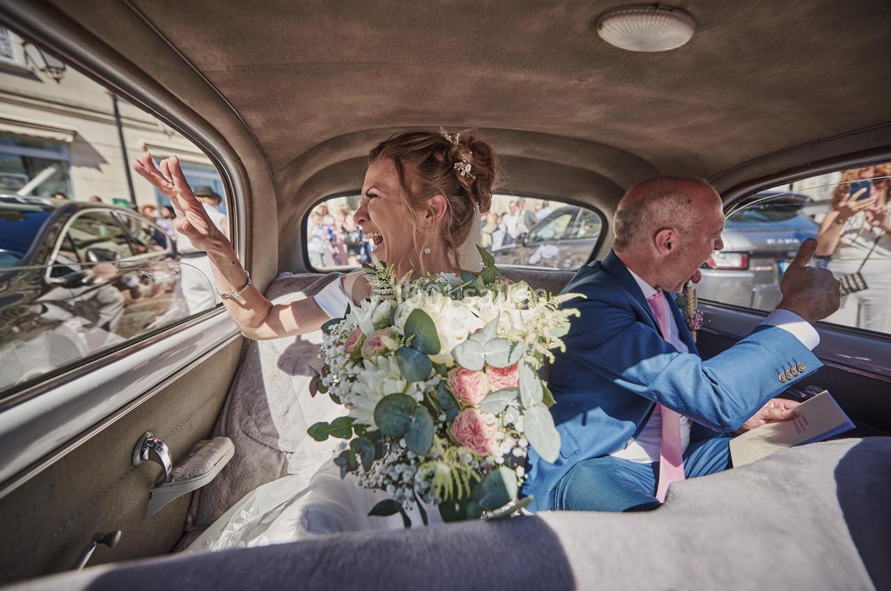 Un couple dans une voiture ancienne salue des invités à travers les fenêtres, un bouquet de mariée au premier plan.