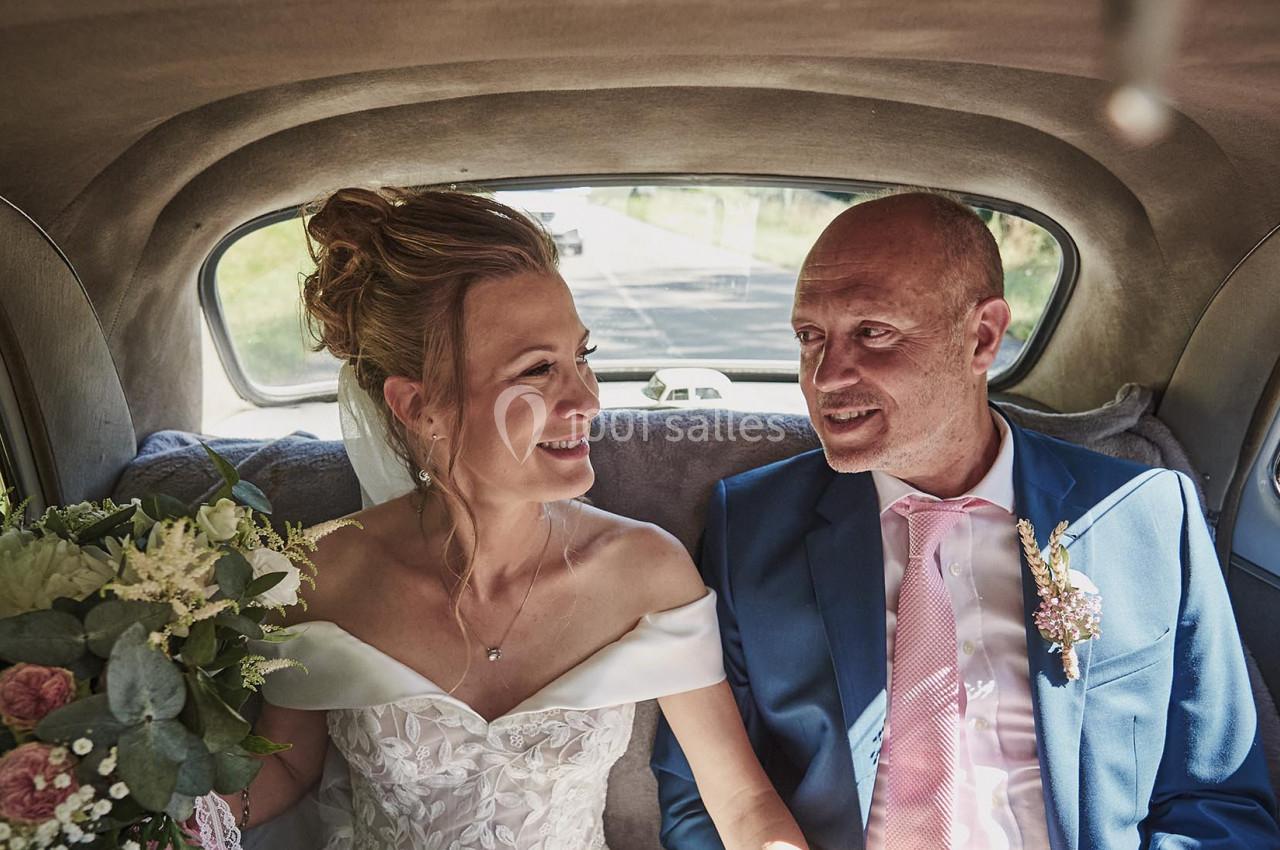 Une mariée en robe blanche et un homme en costume bleu sourient assis à l'arrière d'une voiture ancienne.