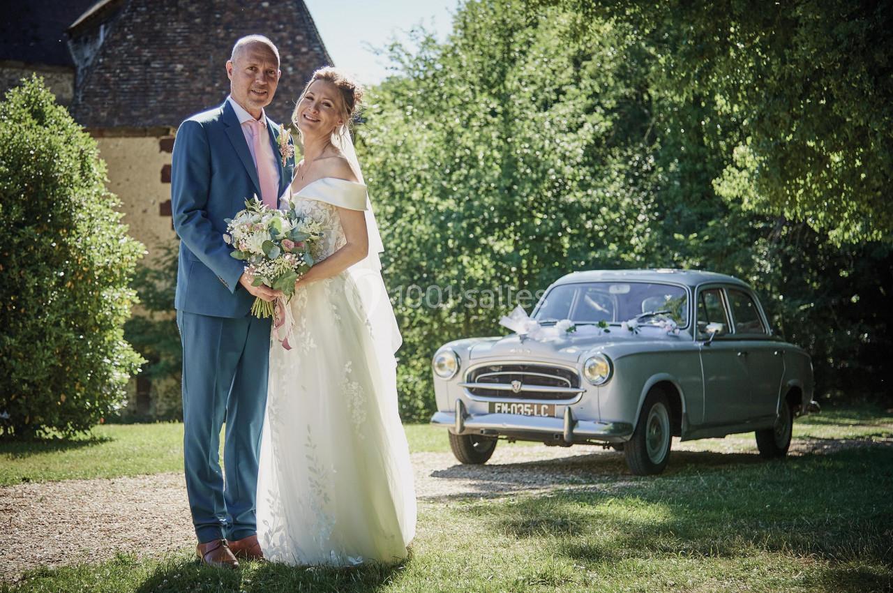 Un couple en tenue de mariage pose devant une voiture ancienne dans un jardin ensoleillé.